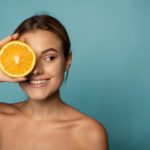caucasian female with clean skin laughing with closed eyes and showing half of fresh orange while advertising benefits of vitamin C